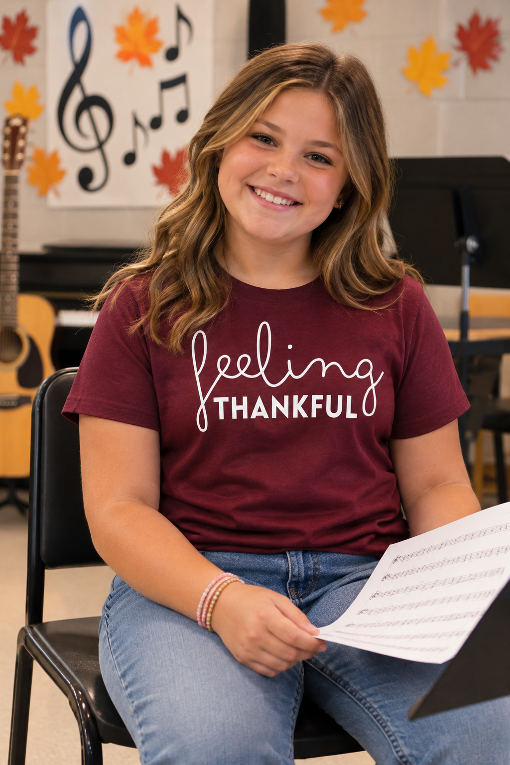 A young girl with long brown hair sits smiling on a black chair, wearing a maroon t-shirt with 'feeling THANKFUL' printed in white script and block letters. She is holding sheet music in a music room setting with musical notes and autumn leaves on the wall.