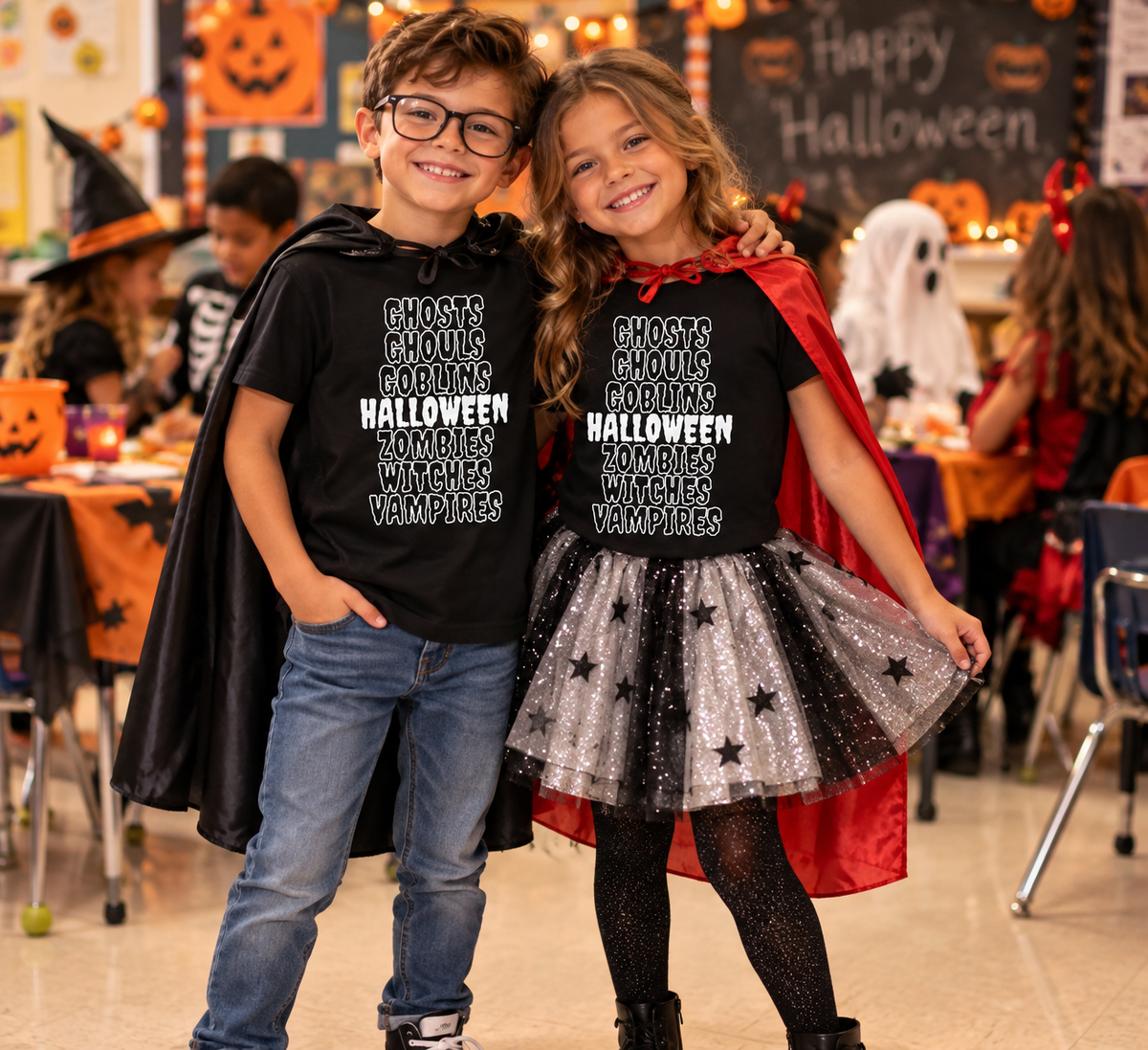 Two children wearing matching black graphic t-shirts with white text, styled as a Halloween costume set. The boy wears a black cape and jeans, while the girl wears a red cape, a sparkly silver tutu skirt with black stars, and black tights.