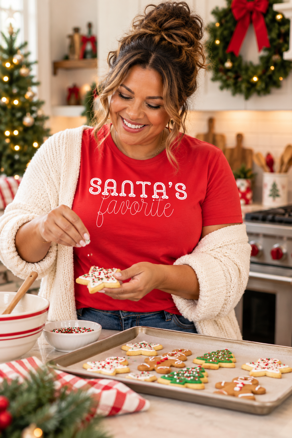 A woman in a cozy kitchen wearing a red graphic t-shirt that reads 'SANTA'S favorite' in white script, sprinkling icing onto decorated holiday cookies on a baking tray.