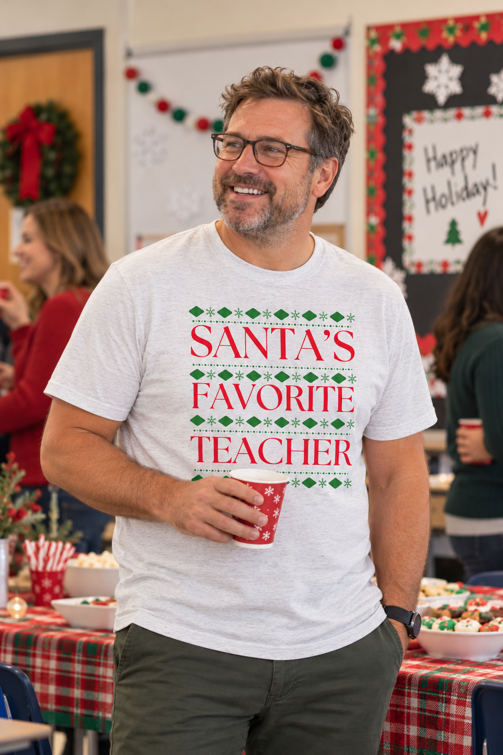 A smiling man wearing glasses and a white graphic T-shirt that reads 'SANTA'S FAVORITE TEACHER' in red with green holiday diamond accents, standing in a decorated holiday classroom or event space.
