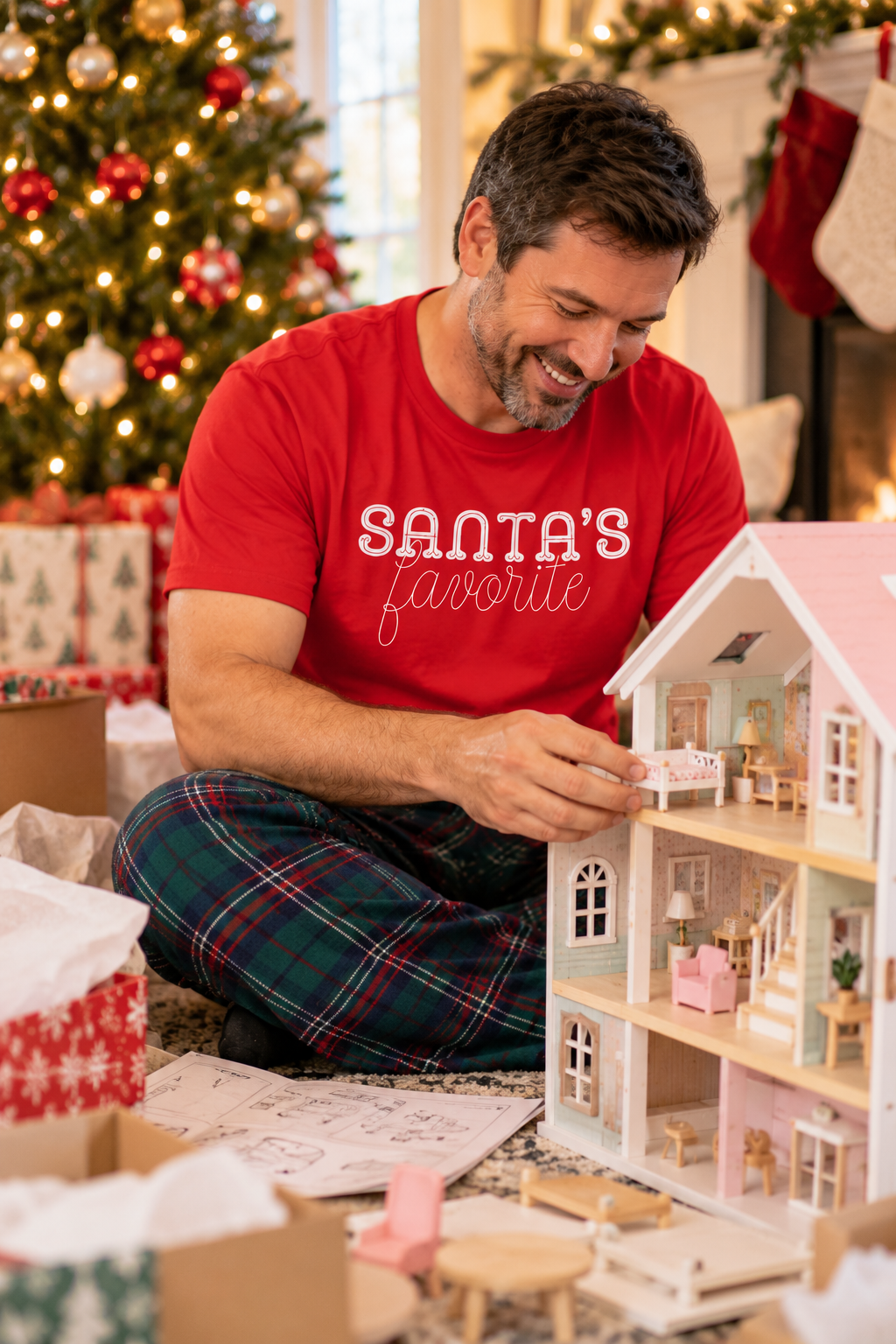 A man in a red t-shirt with 'SANTA'S favorite' printed on it sits on the floor next to a decorated Christmas tree, assembling a miniature dollhouse with tiny furniture pieces.
