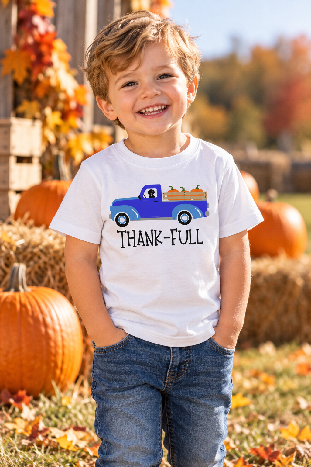 A smiling young boy stands outdoors in a fall yard wearing a white graphic T-shirt with a blue pickup truck and the words 'THANK-FULL' and blue jeans. Pumpkins, hay bales, and autumn leaves are visible in the background.