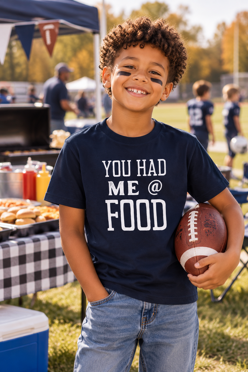 A young boy with curly hair stands outdoors at a food fair, wearing a navy blue graphic t-shirt that reads 'YOU HAD ME @ FOOD' in white text, paired with light blue jeans. He is holding a brown football.
