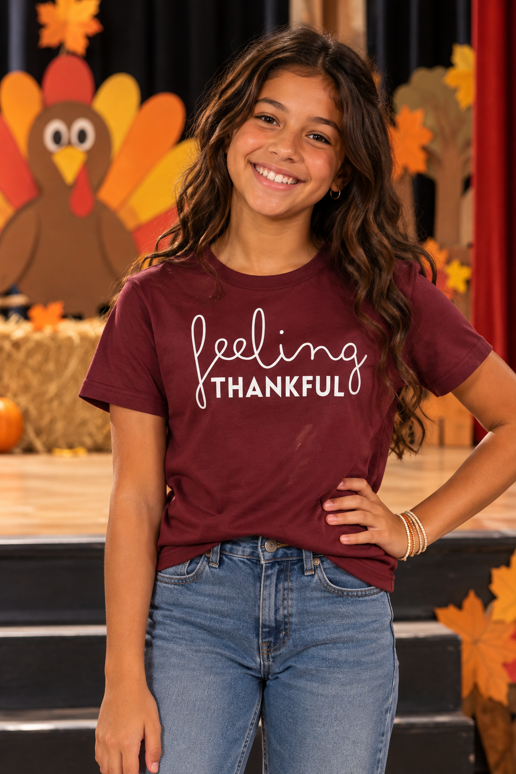 A young girl smiling and posing in a maroon t-shirt with 'feeling THANKFUL' printed in white script and block lettering, paired with blue jeans, set against a festive autumn-themed backdrop.