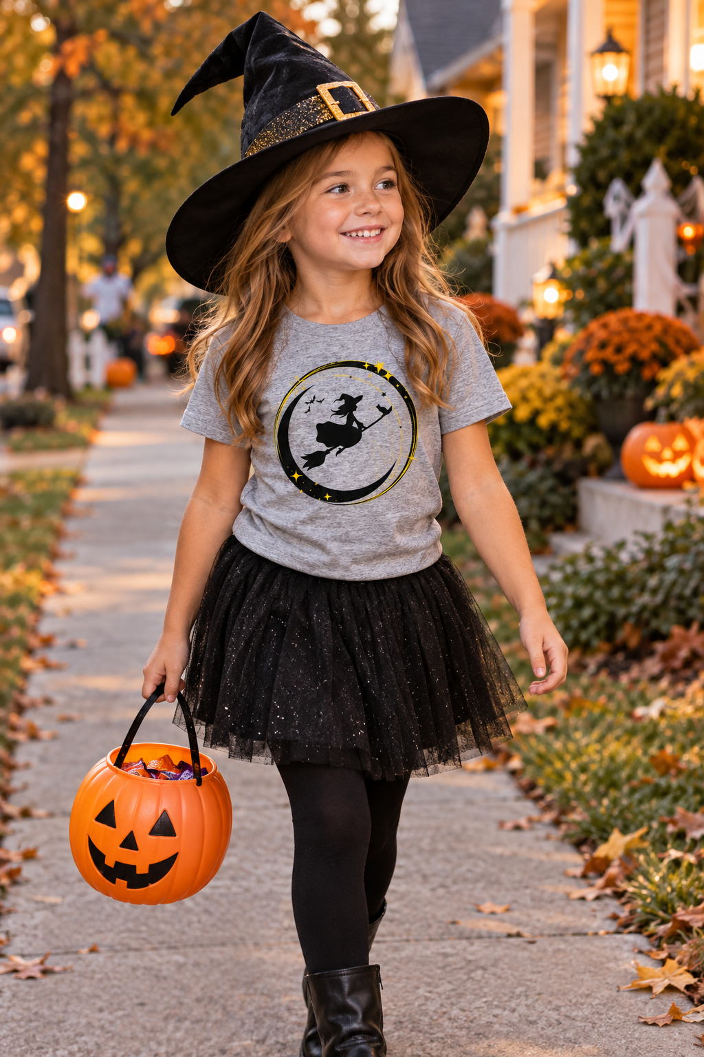 A young girl wearing a witch costume stands on a decorated Halloween street. She is wearing a gray graphic t-shirt with a black and gold witch riding a crescent moon design, a black tulle tutu skirt, black leggings, and a black pointed witch hat.